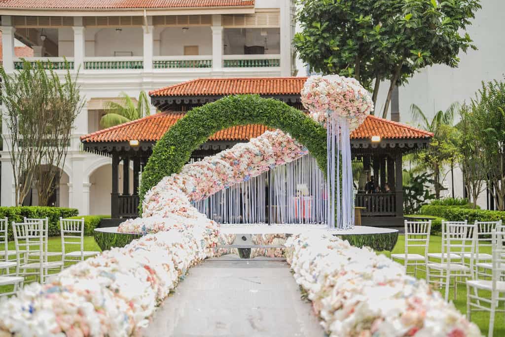 Sculptural outdoor wedding ceremony installation with circular greenery arch, cascading blush and white floral runway and mirror stage platform — Alina & Slava, Raffles Hotel lawn Singapore, floral design by Tomas De Bruyne in collaboration with Create Your Story