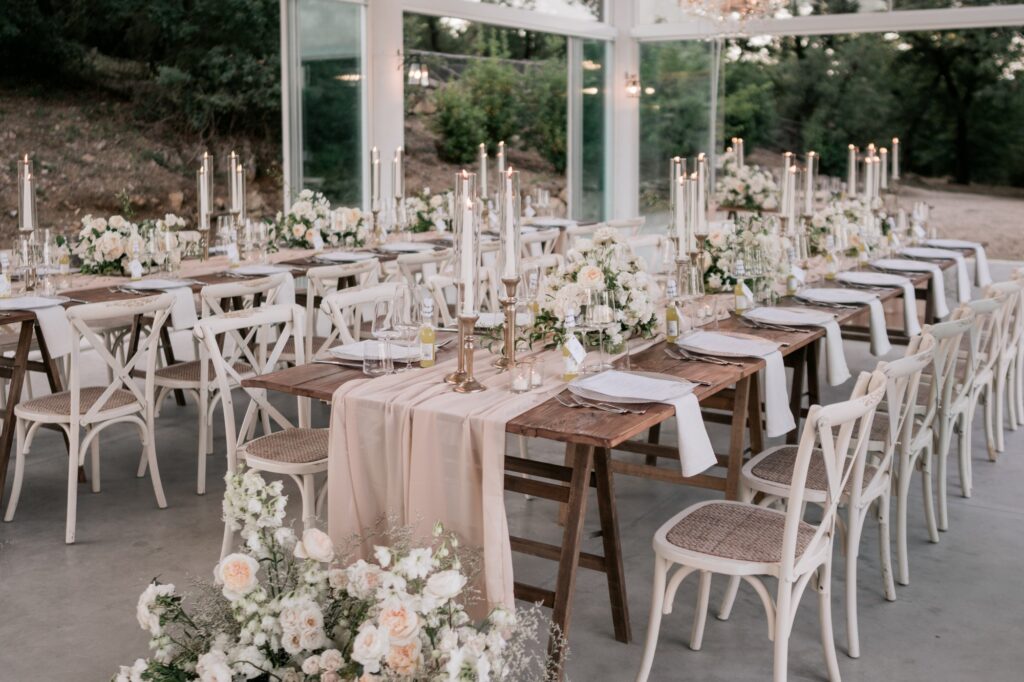 Tuscan glasshouse wedding reception with rustic wooden farm tables, blush linen runners, gold taper candles and low white garden rose and peony centrepieces — Kyller & Parker destination wedding Italy, bespoke floral and styling design by Create Your Story, as featured in British Vogue