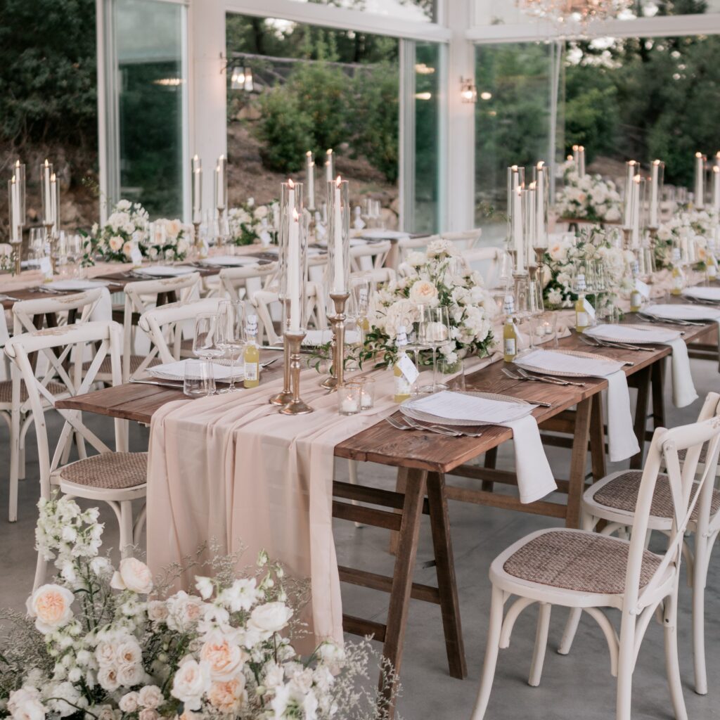Tuscan glasshouse wedding reception with rustic wooden farm tables, blush linen runners, gold taper candles and low white garden rose and peony centrepieces — Kyller & Parker destination wedding Italy, bespoke floral and styling design by Create Your Story, as featured in British Vogue
